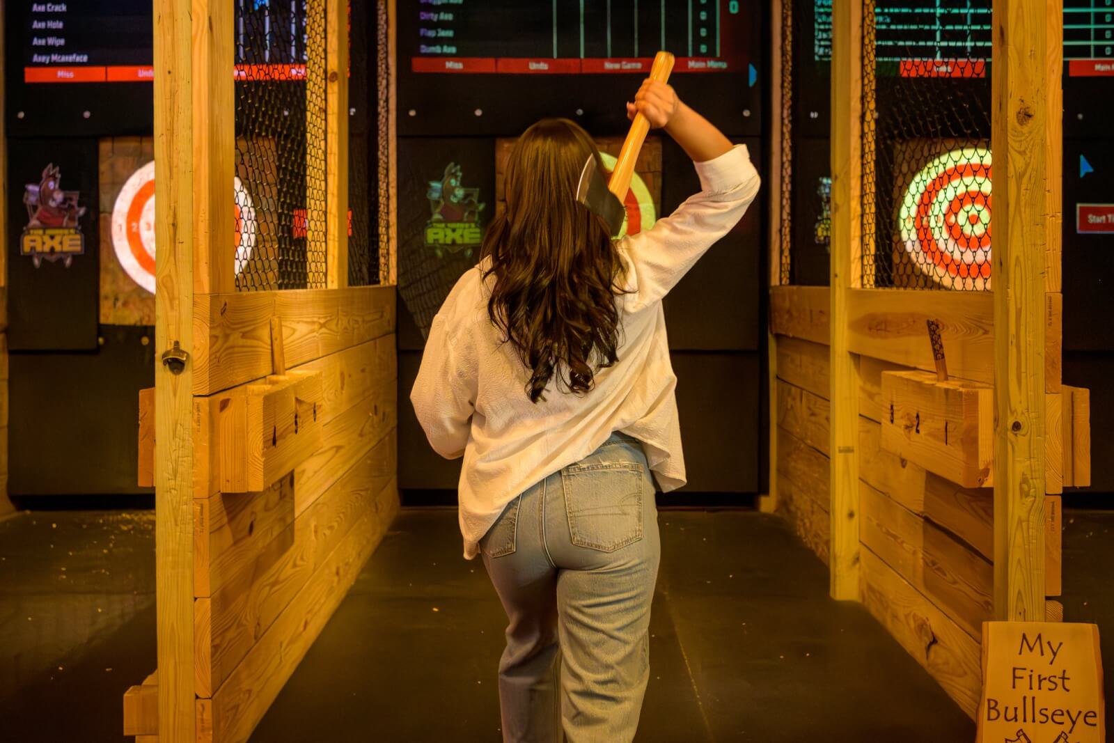 A woman, viewed from behind, has her right arm up with axe in hand preparing to throw it at a target in front of her.